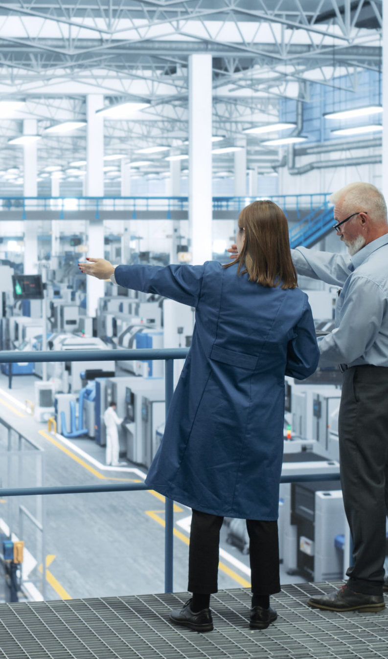 Experienced Male and Female Engineers Standing on a Platform with Their Back to Camera, Using Laptop Computer and Discussing Production at a Modern AI Automated Electronics Manufacture