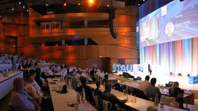 Das Bild zeigt den beeindruckenden Konferenzsaal während des Best Practice Day. Die Teilnehmer sind in mehreren Reihen von Tischen und Stühlen untergebracht, die zur Bühne hin ansteigen. Der Saal ist gut gefüllt, und alle blicken aufmerksam nach vorne, wo Wilhelm Goschy, CEO der STAUFEN.AG, einen Vortrag hält. Sein Name und Bild sind prominent auf einer großen Leinwand hinter ihm zu sehen. Die stimmungsvolle Beleuchtung in warmen Tönen und die farbigen Lichtprojektionen an den Wänden verleihen dem Raum eine einladende und professionelle Atmosphäre. Auf den Tischen der Teilnehmer sind Wasserflaschen und Gläser bereitgestellt, was zur konzentrierten und geschäftigen Stimmung des Events beiträgt.
